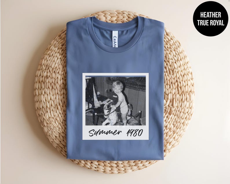 May include: A blue t-shirt with a vintage black and white photo of a child on a toy horse. The photo is in a white frame and has the text "Summer 1980" in a handwritten style. The shirt is folded on a woven mat.