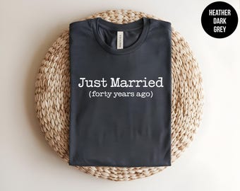 May include: A heather dark grey t-shirt featuring the white text "Just Married (forty years ago)" printed on the front. The shirt has a crew neckline and short sleeves, with a visible brand label inside the collar area.