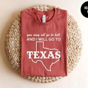 May include: A heather red crew neck t-shirt featuring white text that reads you may all go to hell AND I WILL GO TO TEXAS, with the word TEXAS superimposed over a white outline of the state of Texas. The shirt has a standard short sleeve design.