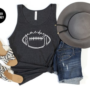 May include: A dark gray tank top with the word "gameday" and a football graphic in white. The image also includes denim shorts, a gray hat, and sandals with striped fabric.