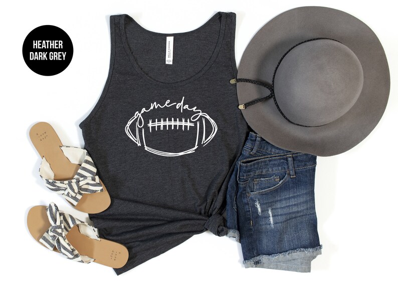 May include: A dark gray tank top with the word "gameday" and a football graphic in white. The image also includes denim shorts, a gray hat, and sandals with striped fabric.