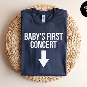 Pode incluir: Uma camiseta azul marinho com o texto "BABY'S FIRST CONCERT" e uma seta branca apontando para baixo.