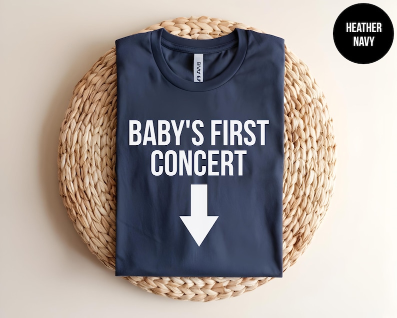 Pode incluir: Uma camiseta azul marinho com o texto "BABY'S FIRST CONCERT" e uma seta branca apontando para baixo.