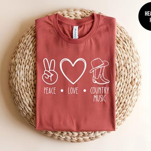 May include: A heather red t-shirt with a white peace sign, heart, cowboy hat, and boots graphic. The text "Peace. Love. Country Music" is printed below the graphic.