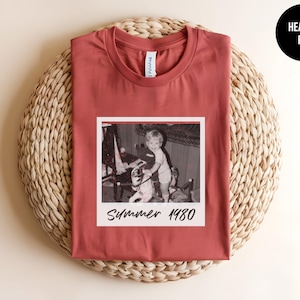May include: Heather red t-shirt featuring a vintage black and white photo with the text "Summer 1980." The shirt has a crew neck and short sleeves. The shirt is displayed on a woven, circular mat.