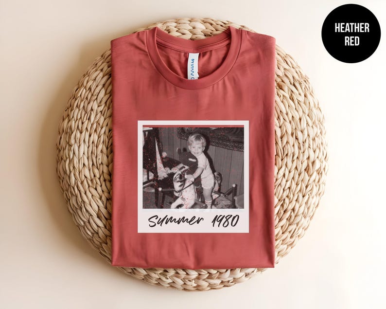 May include: Heather red t-shirt featuring a vintage black and white photo with the text "Summer 1980." The shirt has a crew neck and short sleeves. The shirt is displayed on a woven, circular mat.