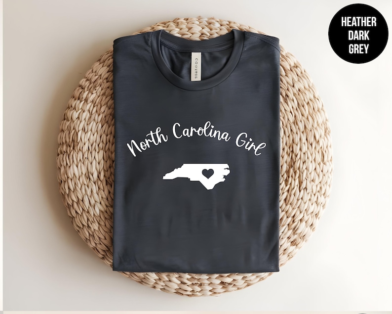 Pu&ograve; includere: T-shirt grigio scuro con la scritta "North Carolina Girl" e un contorno bianco dello stato con un cuore. La maglietta &egrave; piegata su un sottobicchiere circolare intrecciato.