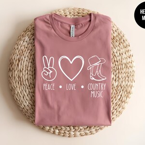 May include: A heather mauve t-shirt with a peace sign, heart, and cowboy boots graphic. The text "Peace. Love. Country Music" is printed below the graphic.