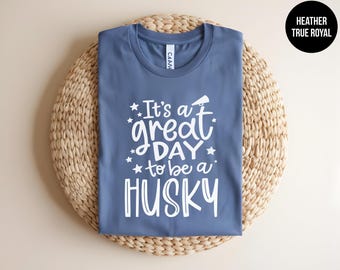 May include: A heather true royal blue t-shirt featuring white screen-printed text that reads It's a great DAY to be a HUSKY, accompanied by small star graphics and a megaphone icon. The shirt has a crew neckline and short sleeves.