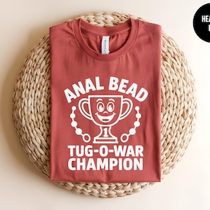 May include: Heather red t-shirt with white text and graphic. The text reads "ANAL BEAD TUG-O-WAR CHAMPION" with a smiling trophy graphic. The shirt is displayed on a woven, circular mat. A black circle with "HEATHER RED" is in the upper right corner.