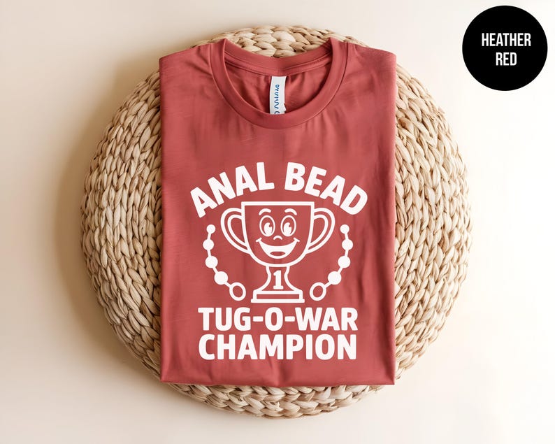 May include: Heather red t-shirt with white text and graphic. The text reads "ANAL BEAD TUG-O-WAR CHAMPION" with a smiling trophy graphic. The shirt is displayed on a woven, circular mat. A black circle with "HEATHER RED" is in the upper right corner.