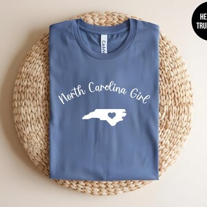 Pu&ograve; includere: Una maglietta blu con la scritta "North Carolina Girl" e un contorno bianco dello stato con un cuore. La maglietta &egrave; piegata su una superficie circolare intrecciata. Il testo "Heather True Royal" &egrave; in un cerchio nero.