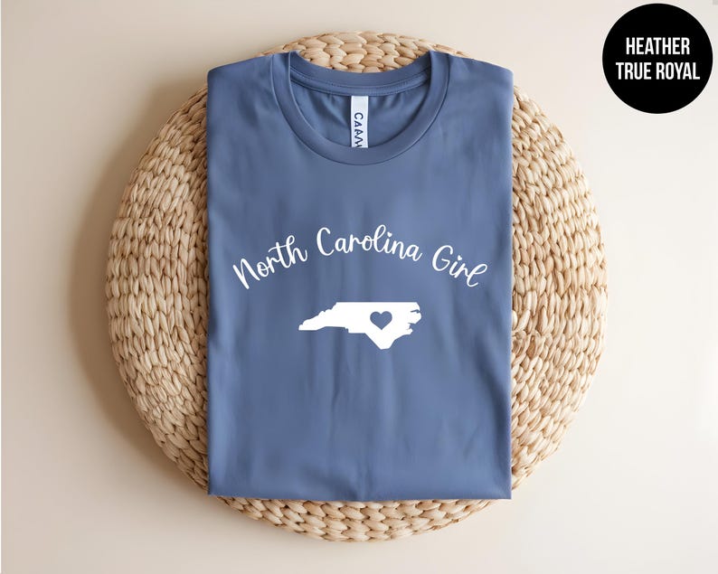Pu&ograve; includere: Una maglietta blu con la scritta "North Carolina Girl" e un contorno bianco dello stato con un cuore. La maglietta &egrave; piegata su una superficie circolare intrecciata. Il testo "Heather True Royal" &egrave; in un cerchio nero.