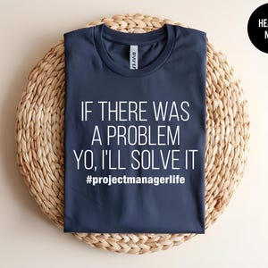 May include: Heather navy t-shirt with the text "IF THERE WAS A PROBLEM YO, I'LL SOLVE IT #projectmanagerlife" in white. The shirt is displayed on a woven, circular placemat.