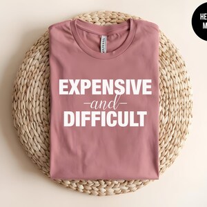 May include: A heather mauve crew neck t-shirt featuring the white text EXPENSIVE and DIFFICULT printed on the front. The shirt has short sleeves and a standard fit, with the text displayed in a bold, sans-serif font for the main words.