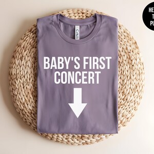 Pode incluir: Camiseta roxa em helanca com o texto "BABY'S FIRST CONCERT" e uma seta branca apontando para baixo.