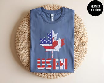 May include: A heather true royal blue t-shirt featuring a distressed graphic of a maple leaf split between the American flag and the Canadian flag. Below the leaf, the text USEH is printed, with the letters US in American flag colors and EH in Canadian flag colors.
