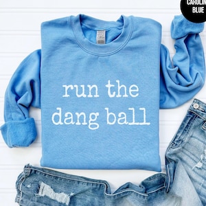 May include: A Carolina blue sweatshirt with the phrase "run the dang ball" in white block letters. The sweatshirt is folded with a pair of blue jeans. The sleeves are cuffed.