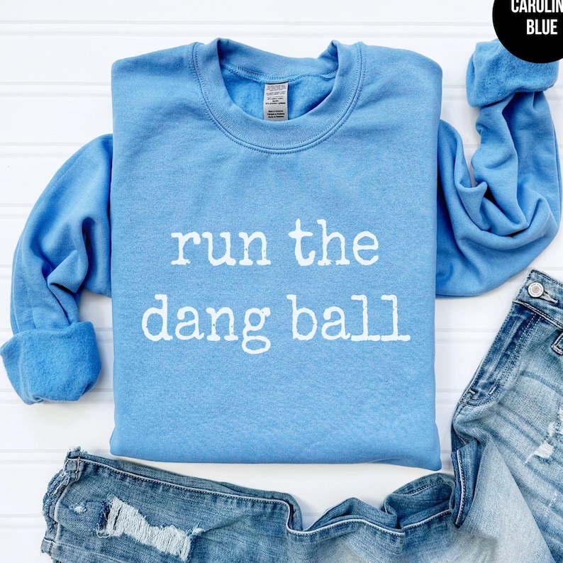 May include: A Carolina blue sweatshirt with the phrase "run the dang ball" in white block letters. The sweatshirt is folded with a pair of blue jeans. The sleeves are cuffed.