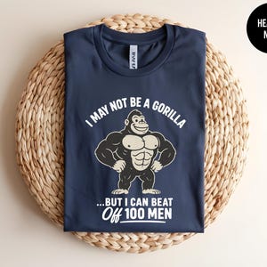 May include: A heather navy blue t-shirt featuring a graphic of a muscular gorilla with the white text "I MAY NOT BE A GORILLA ...BUT I CAN BEAT OFF 100 MEN". The shirt has a crew neckline and short sleeves, with a small black circular label in the corner.