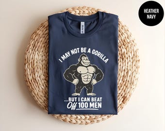 May include: A heather navy blue t-shirt featuring a graphic of a muscular gorilla with the white text "I MAY NOT BE A GORILLA ...BUT I CAN BEAT OFF 100 MEN". The shirt has a crew neckline and short sleeves, with a small black circular label in the corner.