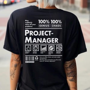 May include: Black t-shirt with white text that reads "PROJECT-MANAGER." The shirt includes icons and phrases like "Requires Coffee," "Always on Time," and "Fluent in Excel." The text also states "100% Genius 100% Chaos."