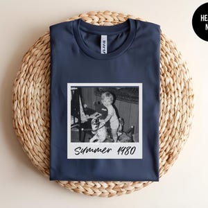 May include: Navy blue t-shirt featuring a black and white photo print of a child with a toy. The photo is in a white frame with the text "Summer 1980". The shirt is displayed on a woven, circular surface.