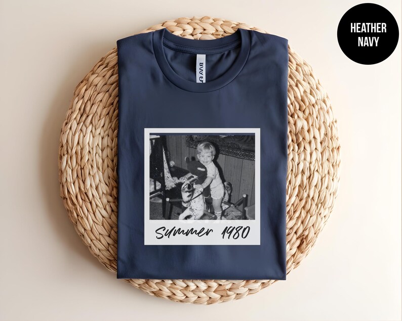 May include: Navy blue t-shirt featuring a black and white photo print of a child with a toy. The photo is in a white frame with the text "Summer 1980". The shirt is displayed on a woven, circular surface.