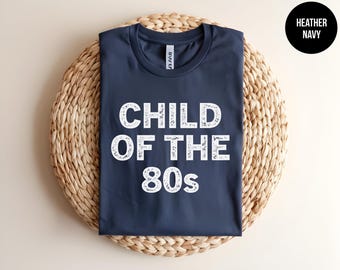 May include: A heather navy blue crew neck t-shirt featuring the white distressed text "CHILD OF THE 80s" printed on the front. The shirt has a standard short-sleeve design and a visible brand label inside the collar area.