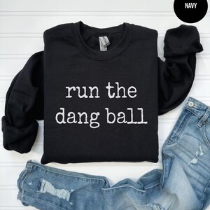 May include: A navy blue sweatshirt with the phrase "run the dang ball" printed in white. The sweatshirt is displayed with a pair of distressed blue jeans. The word "NAVY" is in a black circle in the upper right corner.