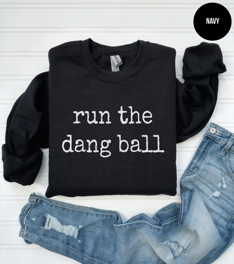 May include: A navy blue sweatshirt with the phrase "run the dang ball" printed in white. The sweatshirt is displayed with a pair of distressed blue jeans. The word "NAVY" is in a black circle in the upper right corner.