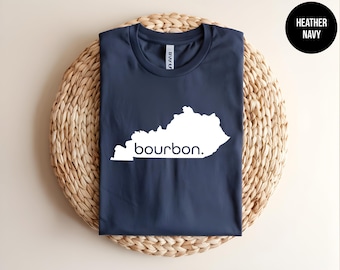 May include: A heather navy blue t-shirt featuring a white graphic outline of the state of Kentucky with the word bourbon. printed in lowercase white letters across the center of the state shape. The shirt has a crew neckline and short sleeves.