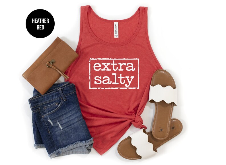 May include: A red tank top with the text 'extra salty' in white. The tank top is displayed on a white background with a brown clutch, blue denim shorts, and white sandals.
