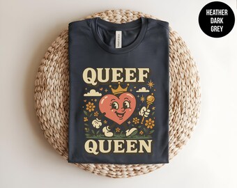 May include: A heather dark grey t-shirt featuring a graphic of a smiling heart character wearing a crown and holding a scepter. The text "QUEEF QUEEN" is printed in a bold, cream-colored font above and below the heart, surrounded by small flowers, clouds, and stars.