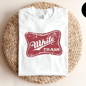 Peut inclure: Un t-shirt blanc à manches courtes avec un motif rouge vieilli sur la poitrine. Le motif est une forme rectangulaire aux coins arrondis contenant le texte White TRASH dans une police blanche stylisée. Le t-shirt a un col rond.