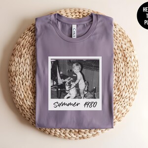 May include: A heather purple t-shirt featuring a black and white photo with the text "Summer 1980." The shirt has a crew neck and is displayed on a woven, circular surface. A black circle with the text "Heather Team Purple" is in the upper right corner.