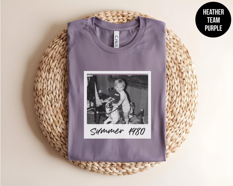 May include: A heather purple t-shirt featuring a black and white photo with the text "Summer 1980." The shirt has a crew neck and is displayed on a woven, circular surface. A black circle with the text "Heather Team Purple" is in the upper right corner.