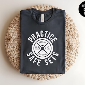 May include: A heather dark grey t-shirt featuring a white graphic of a barbell weight plate. The text reads "PRACTICE SAFE SETS" in a circular arrangement around the weight, which is labelled "BARBELL", "STANDARD", "20.4 KGS", and "45 LBS".