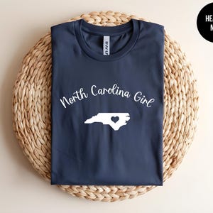 Pu&ograve; includere: Una maglietta blu navy con le parole "North Carolina Girl" in corsivo bianco sopra un contorno bianco della Carolina del Nord con un cuore. La maglietta &egrave; esposta su un tappetino circolare intrecciato. Un cerchio nero con "Heather Navy" &egrave; in alto a destra.