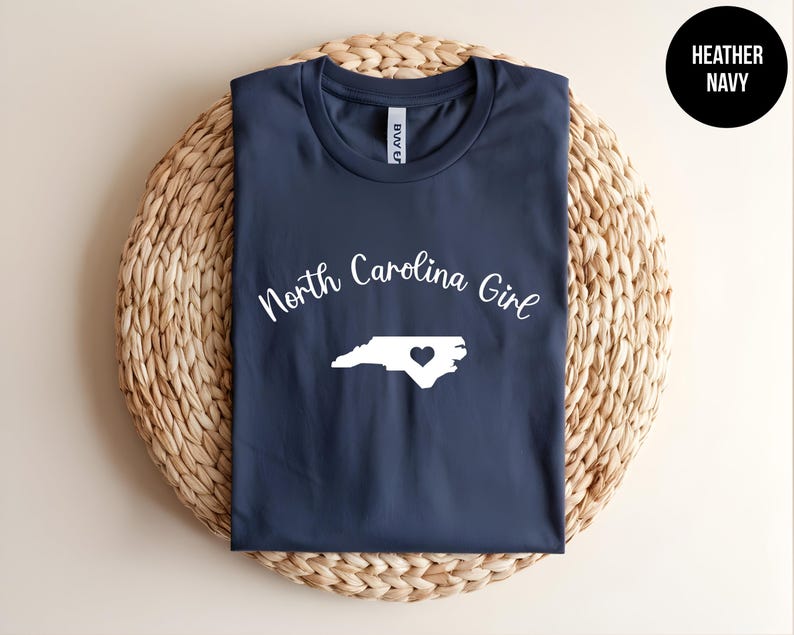 Pu&ograve; includere: Una maglietta blu navy con le parole "North Carolina Girl" in corsivo bianco sopra un contorno bianco della Carolina del Nord con un cuore. La maglietta &egrave; esposta su un tappetino circolare intrecciato. Un cerchio nero con "Heather Navy" &egrave; in alto a destra.