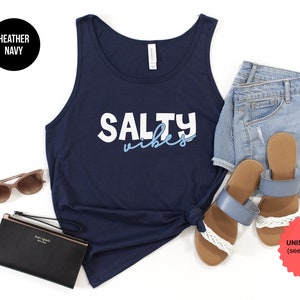 May include: A navy blue tank top with the text "Salty Vibes" in white and light blue lettering. The tank top is laying flat on a white background with a pair of blue sandals, denim shorts, sunglasses, and a black wallet.