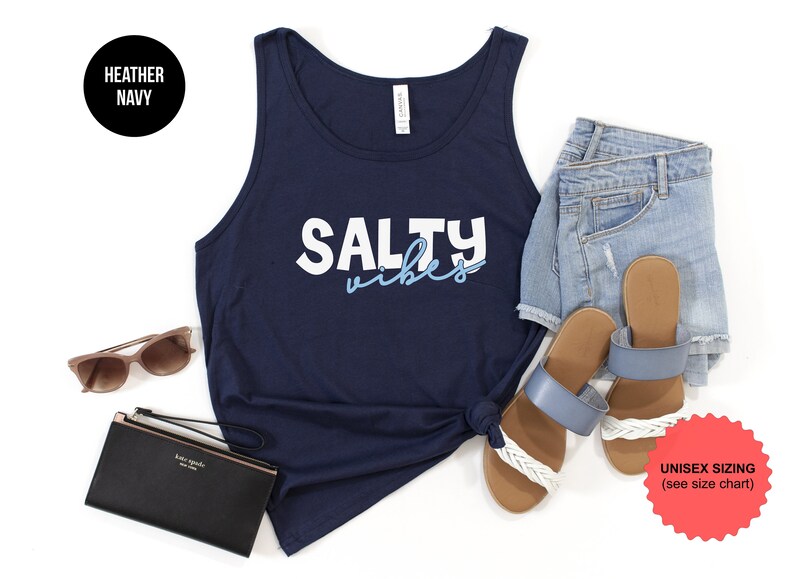 May include: A navy blue tank top with the text "Salty Vibes" in white and light blue lettering. The tank top is laying flat on a white background with a pair of blue sandals, denim shorts, sunglasses, and a black wallet.