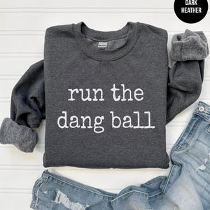 May include: Dark heather gray sweatshirt with the phrase "run the dang ball" in white lettering. The sweatshirt has a crew neck and long sleeves. Faded blue denim jeans are visible at the bottom of the image.