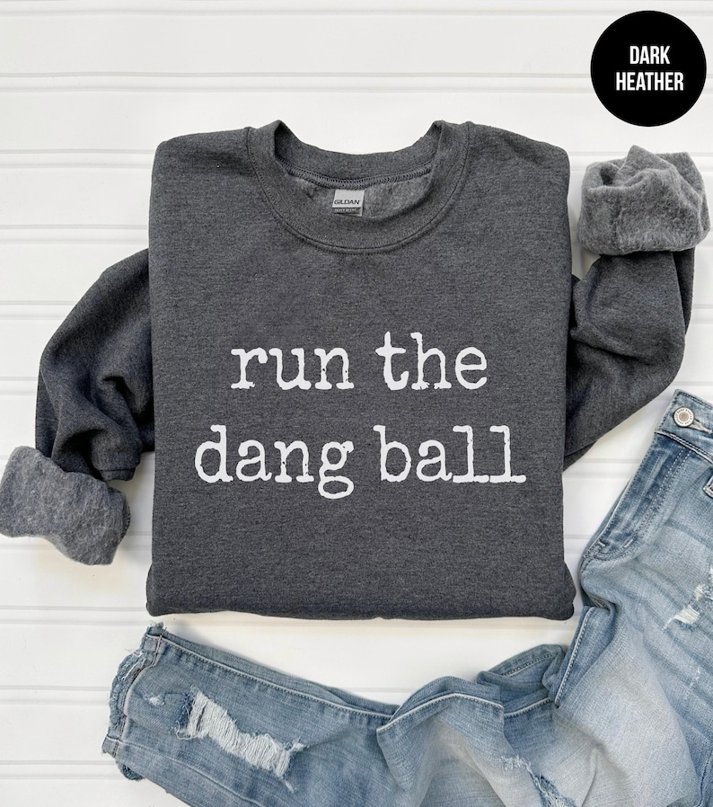 May include: Dark heather gray sweatshirt with the phrase "run the dang ball" in white lettering. The sweatshirt has a crew neck and long sleeves. Faded blue denim jeans are visible at the bottom of the image.