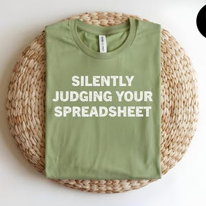 May include: A heather green t-shirt with a crew neckline featuring the white text "SILENTLY JUDGING YOUR SPREADSHEET" printed in a bold, sans-serif font across the center of the chest. The garment has short sleeves and a standard fit.