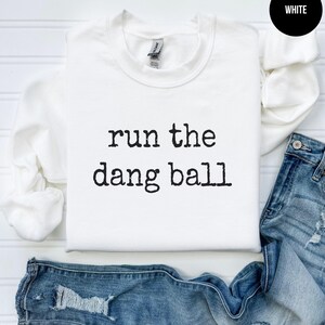 May include: A white crewneck sweatshirt with the phrase "run the dang ball" printed in black. The sweatshirt is displayed with a pair of blue jeans. A black circle with the word "WHITE" is in the upper right corner.