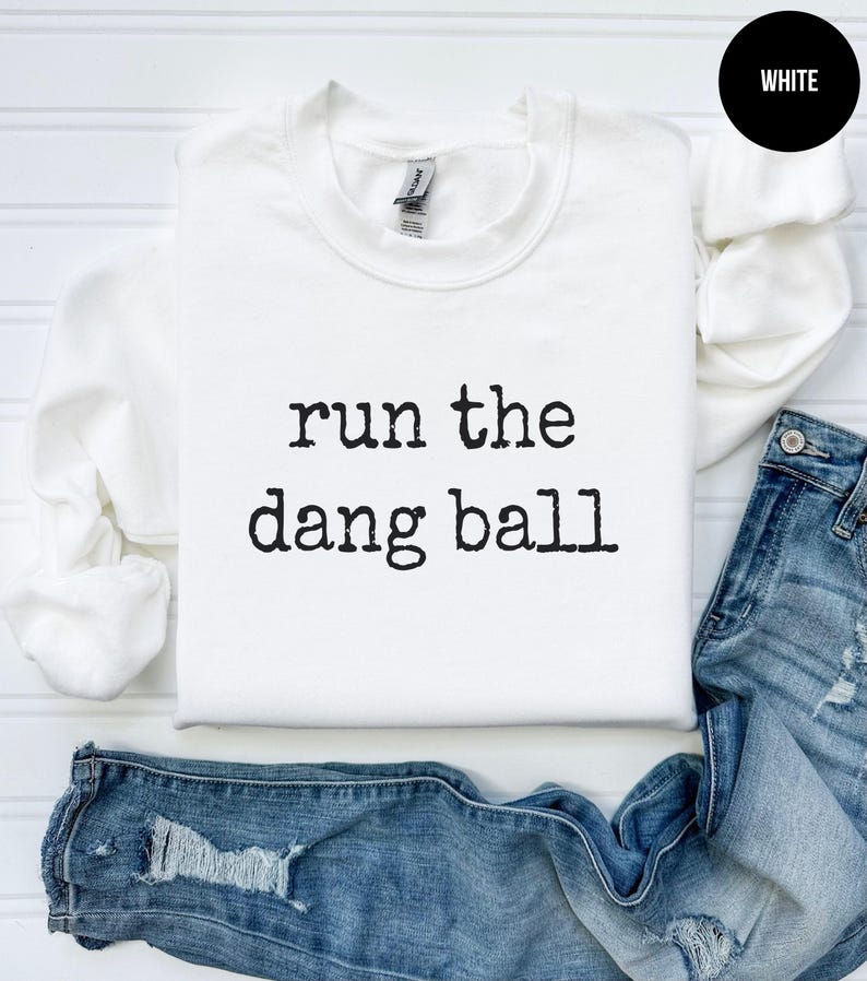 May include: A white crewneck sweatshirt with the phrase "run the dang ball" printed in black. The sweatshirt is displayed with a pair of blue jeans. A black circle with the word "WHITE" is in the upper right corner.