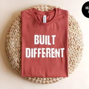 May include: A heather red short-sleeved t-shirt with a crew neckline. The front of the shirt features the text "BUILT DIFFERENT" printed in a distressed white font. The garment has a standard fit and a smooth, solid-colored fabric texture.