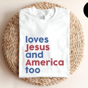 May include: A white short-sleeved t-shirt featuring the text "loves Jesus and America too" printed in alternating blue and red sans-serif lettering. The shirt has a crew neckline and is displayed folded flat with a white color label in the corner.
