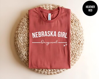 May include: A heather red crew neck t-shirt featuring the white text NEBRASKA GIRL in a bold sans-serif font, followed by the word Original in a white script font, a horizontal line, and two small white heart icons on the right side.